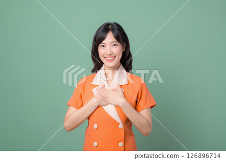 Southeast asian woman smiling with hands on chest in orange dress standing against green background showing gratitude Southeast asian woman smiling with hands on chest in orange dress standing against green background showing gratitude 126896714