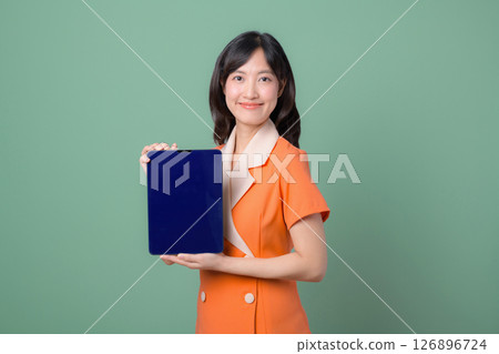Southeast asian woman in orange suit holding blank blue tablet with cheerful expression on green background Southeast asian woman in orange suit holding blank blue tablet with cheerful expression on green background 126896724