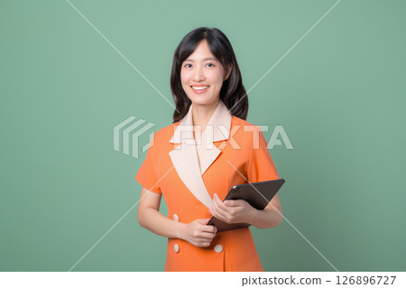 Southeast asian woman in orange dress holding tablet smiling confidently against green background, representing modern professional and positive attitude in business or education 126896727