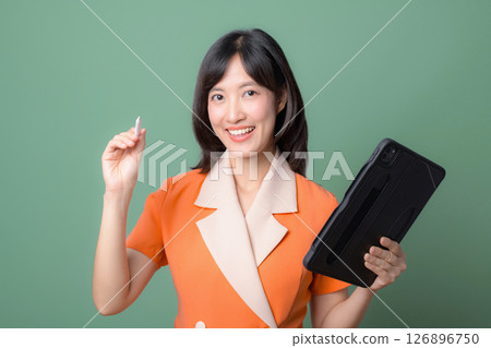 Southeast asian woman in orange suit smiling holding tablet and stylus pen on green background showing cheerful expression Southeast asian woman in orange suit smiling holding tablet and stylus pen on green background showing cheerful expression 126896750