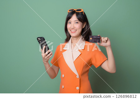Southeast asian woman in orange dress smiling holding smartphone and credit card against green background showing cheerful expression 126896812