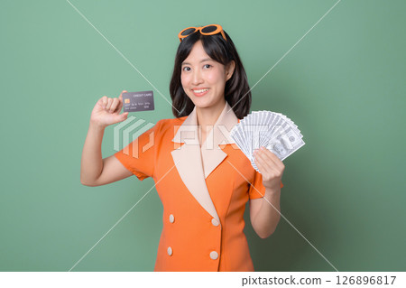 Southeast asian woman in orange dress holding credit card and cash smiling against green background financial success concept Southeast asian woman in orange dress holding credit card and cash smiling against green background financial success concept 126896817