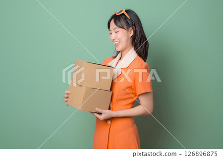 Southeast asian woman in orange dress smiling while holding two cardboard boxes against green background Southeast asian woman in orange dress smiling while holding two cardboard boxes against green background 126896875