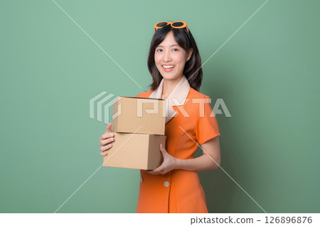 Southeast asian woman in orange dress holding cardboard boxes smiling against green background cheerful delivery concept Southeast asian woman in orange dress holding cardboard boxes smiling against green background cheerful delivery concept 126896876