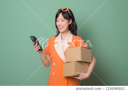 Southeast asian woman in orange dress holding cardboard boxes and using smartphone standing against green background smiling with cheerful expression Southeast asian woman in orange dress holding cardboard boxes and using smartphone standing against green background smiling with cheerful expression 126896881