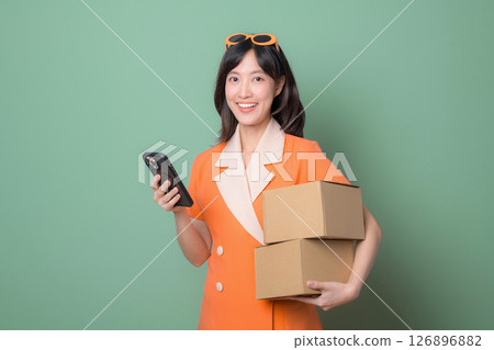 Southeast asian woman holding packages and using smartphone for internet online shopping, smiling and wearing orange dress with sunglasses against green background Southeast asian woman holding packages and using smartphone for internet online shopping, smiling and wearing orange dress with sunglasses against green background 126896882