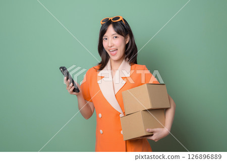 Happy woman in orange dress holds cardboard boxes and phone against green background, delivery and e commerce concept 126896889