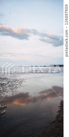 Spring landscape. Panoramic view of beautiful dawn on bay. Cumulus clouds over water in bright light. Ice, snow and rocks on coastline. Rising rays of sun are reflected in sea. Vertical photo. 126897609