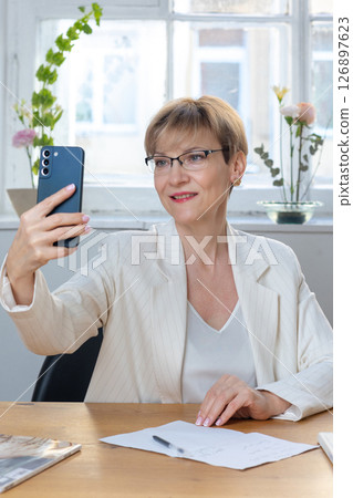 Close-up portrait happy woman in business suit at her desk. Female holding mobile phone, taking selfie with smartphone posing on front camera. Vertical photo. High quality photo Close-up portrait happy woman in business suit at her desk. Female holding mobile phone, taking selfie with smartphone posing on front camera. Vertical photo. High quality photo 126897623