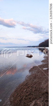 Spring landscape. Panoramic view of beautiful dawn on bay. Cumulus clouds over water in bright light. Ice, snow and rocks on coastline. Rising rays of sun are reflected in sea. Vertical photo. Spring landscape. Panoramic view of beautiful dawn on bay. Cumulus clouds over water in bright light. Ice, snow and rocks on coastline. Rising rays of sun are reflected in sea. Vertical photo. 126897696