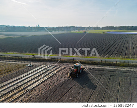 A tractor works on a large agricultural field near a road, with neat rows of soil stretching into the distance. Morning sun casts soft light across the landscape. A tractor works on a large agricultural field near a road, with neat rows of soil stretching into the distance. Morning sun casts soft light across the landscape. 126898247