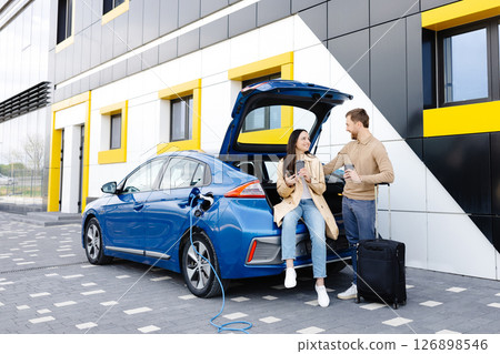 Young friends charging automobile battery from public station and standing near parking for electric car. Man and woman talking and drinking coffee in city Young friends charging automobile battery from public station and standing near parking for electric car. Man and woman talking and drinking coffee in city 126898546