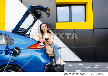 Young woman waiting charging automobile battery from small public station and using smartphone while charging automobile Young woman waiting charging automobile battery from small public station and using smartphone while charging automobile 126898560