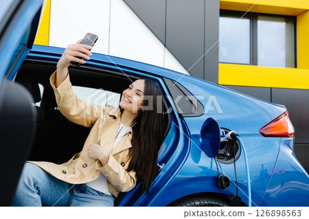 Business lady sitting inside charging electric car and using modern smartphone. Technology and transport concept. Business lady sitting inside charging electric car and using modern smartphone. Technology and transport concept. 126898563