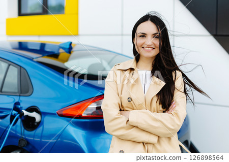 Woman is standing at the car and charging electric car at the charging station 126898564
