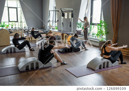 Group of women doing exercises on back spine corrector. Reformer pilates studio machine for fitness workouts in gym. Fit, healthy and strong authentical body. Fitness concept. 126899109
