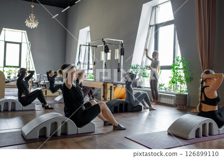 Group of women doing exercises on back spine corrector. Reformer pilates studio machine for fitness workouts in gym. Fit, healthy and strong authentical body. Fitness concept. Group of women doing exercises on back spine corrector. Reformer pilates studio machine for fitness workouts in gym. Fit, healthy and strong authentical body. Fitness concept. 126899110