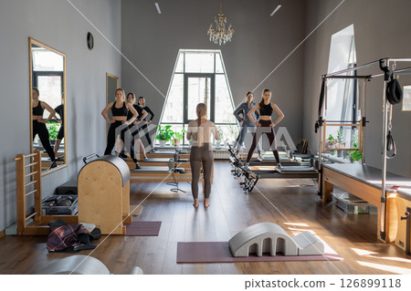 Young caucasian women exercising on pilates reformers in a bright studio. Their workout focuses on core strength and flexibility in a modern fitness environment. Young caucasian women exercising on pilates reformers in a bright studio. Their workout focuses on core strength and flexibility in a modern fitness environment. 126899118