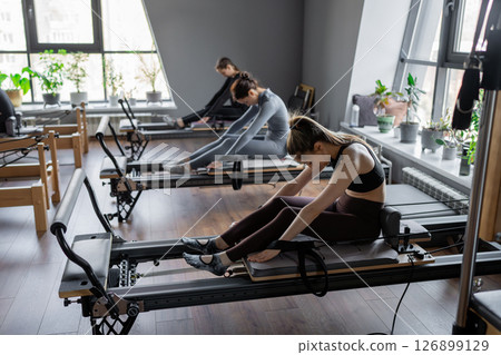 Group of caucasian women exercising with trainer on pilates reformers in a bright studio. Their workout focuses on core strength and flexibility in a modern fitness environment Group of caucasian women exercising with trainer on pilates reformers in a bright studio. Their workout focuses on core strength and flexibility in a modern fitness environment 126899129