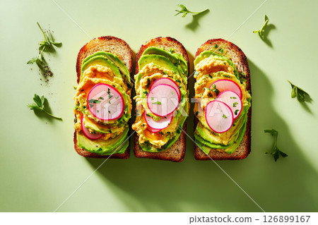 Three beautifully arranged breakfast toasts topped with avocado slices, chickpeas, radish, and cilantro are displayed on a bright green plate, showcasing a colorful and healthy morning meal 126899167