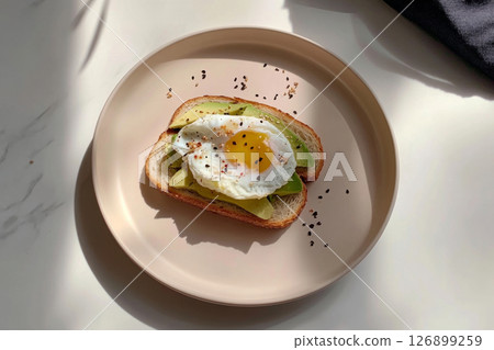 A beautifully arranged breakfast plate features toasted bread topped with a perfectly fried egg. Fresh avocado slices and a lemon half complement the hearty meal in a warm setting 126899259