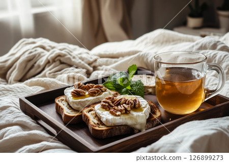 Deliciously crafted toasts topped with cream cheese, pecans, and honey placed on a wooden tray beside a cup of tea, all set on a soft blanket in a serene bedroom Deliciously crafted toasts topped with cream cheese, pecans, and honey placed on a wooden tray beside a cup of tea, all set on a soft blanket in a serene bedroom 126899273