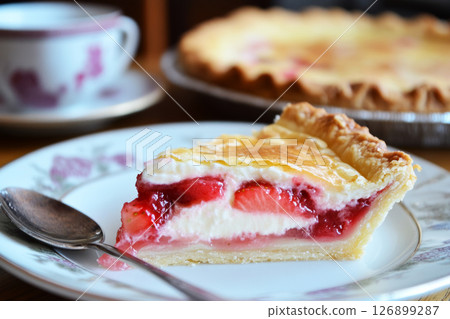 A slice of strawberry tart rests on an elegant floral plate, topped with fresh strawberries and glossy glaze. A silver spoon lies beside it, inviting a sweet indulgence 126899287