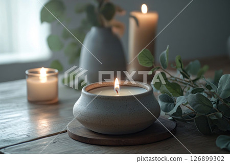 A ceramic vase holds a flickering candle accompanied by a sprig of eucalyptus. Soft natural light filters through sheer curtains, creating a tranquil atmosphere A ceramic vase holds a flickering candle accompanied by a sprig of eucalyptus. Soft natural light filters through sheer curtains, creating a tranquil atmosphere 126899302