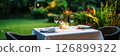 A beautifully arranged dining table in a vibrant garden features fine tableware and glasses, creating a serene atmosphere for guests to enjoy a meal outdoors, banner 126899322