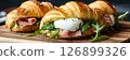 A beautifully plated croissant sandwich with a runny poached egg, savory ham, and fresh arugula, set against a bright background. A delicious and elegant morning meal with French inspiration, banner 126899326