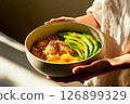 A person holds a bowl containing quinoa, sliced avocado, and fresh mango. The arrangement showcases vibrant colors, enhanced by warm sunlight 126899329