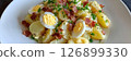 A hearty serving of Kartoffelsalat, adorned with sliced hard-boiled eggs and crispy bacon, showcases a traditional German dish served in a casual dining environment, banner 126899330