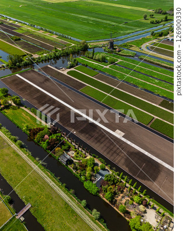 Aerial view of Dutch farmland with green fields, canals, and a traditional windmill. Neatly divided plots and vibrant vegetation highlight the beauty of rural agricultural landscape. Aerial view of Dutch farmland with green fields, canals, and a traditional windmill. Neatly divided plots and vibrant vegetation highlight the beauty of rural agricultural landscape. 126900330