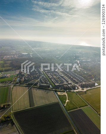 Hazy aerial view of a Dutch town merging into farmland, with the North Sea shimmering on the horizon. A mix of urban housing, fields, and forest under soft morning light. Hazy aerial view of a Dutch town merging into farmland, with the North Sea shimmering on the horizon. A mix of urban housing, fields, and forest under soft morning light. 126900334