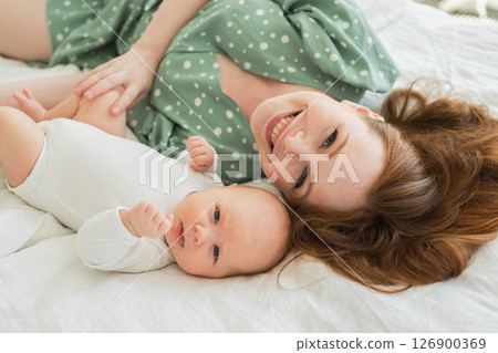 Happy family at home. Mother playing with little toddler child on bed. Mom baby relax playing having fun together. Mother looks at camera with love care. Mom of breast feeding baby rest in living room Happy family at home. Mother playing with little toddler child on bed. Mom baby relax playing having fun together. Mother looks at camera with love care. Mom of breast feeding baby rest in living room 126900369