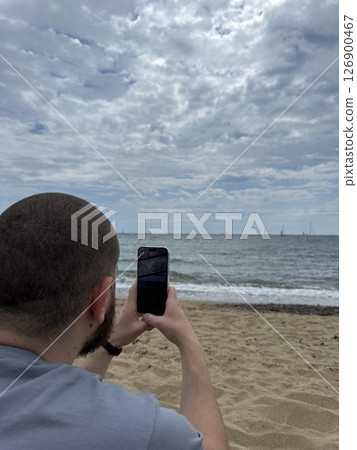 Guy, tourist, takes photos of the sea on his phone sitting on the shore, vertical photo 126900467