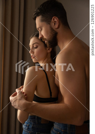 Shirtless man in jeans embracing woman in black top and jeans standing closely in intimate moment Shirtless man in jeans embracing woman in black top and jeans standing closely in intimate moment 126901225