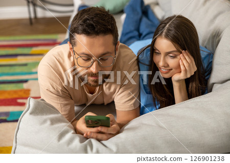Happy young woman and man using smartphone together lying on couch Happy young woman and man using smartphone together lying on couch 126901238