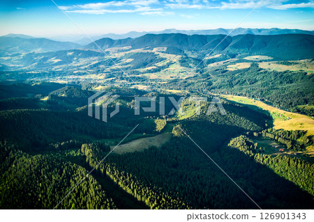 Aerial view of densely forested mountains stretching into distance under clear blue sky with scattered clouds. Landscape reveals valleys and distant fields, showcasing natural beauty of region. Aerial view of densely forested mountains stretching into distance under clear blue sky with scattered clouds. Landscape reveals valleys and distant fields, showcasing natural beauty of region. 126901343
