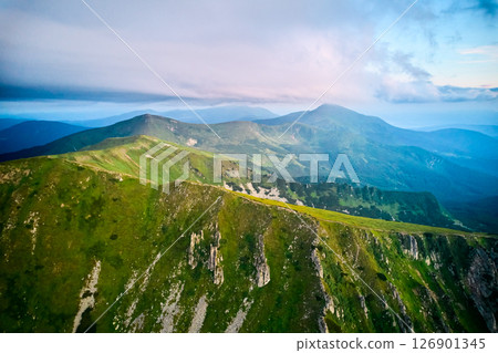 Breathtaking aerial view of green, rugged mountain peaks under dramatic sky. Scene captures contrast between vibrant landscape and moody clouds, with sunlight breaking through. Carpathians, Chornogora 126901345
