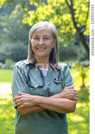 Caucasian female nurse standing with her arms crossed on a greenery background Caucasian female nurse standing with her arms crossed on a greenery background 126901591