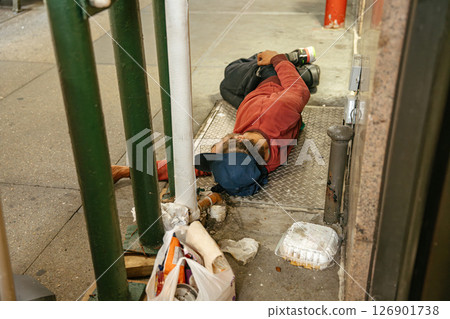 A man in a red sweatshirt sleeps on the ground beside trash on a New York street. Urban poverty and homelessness are visible in the city environment. 126901738