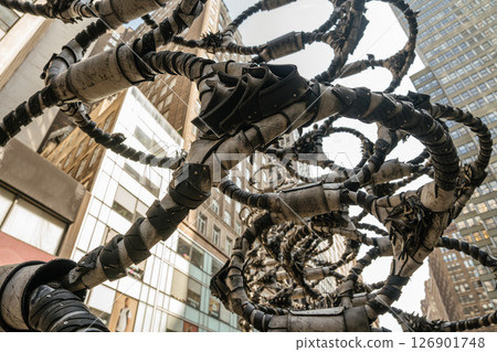 A dramatic close-up of a twisted metal sculpture in New York showcases intricate armored spirals. The background includes glass buildings and urban reflections in Midtown Manhattan. 126901748