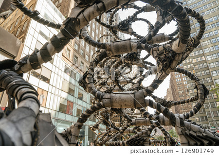 Abstract metal sculpture spirals upward between NYC buildings. It features twisting coils and layered textures. 126901749