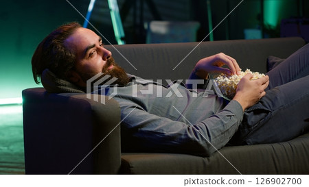 Amused man lounged on couch laughing at funny comedy movie while eating from bowl of popcorn. Cheerful person laying down in apartment amused by hilarious humor in film scene, camera A close up Amused man lounged on couch laughing at funny comedy movie while eating from bowl of popcorn. Cheerful person laying down in apartment amused by hilarious humor in film scene, camera A close up 126902700