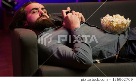 Gamer lounged on couch engaging in multiplayer game using gamepad, eating snack. Person in home theatre playing videogames on gaming console with bowl of popcorn in lap, camera A 126902709