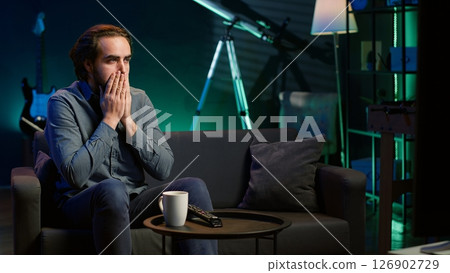 Man at home shocked by thriller film on TV, drinking coffee. Stupefied person at home left with eyes wide open by dramatic plot twist, watching suspenseful movie on TV, enjoying beverage, camera A 126902729