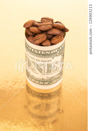 A single dollar bill, rolled and stuffed with coffee beans, rests on a gold surface with loose beans 126903213