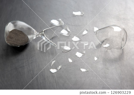 A broken hourglass with dark sand spilling out on a metallic surface A broken hourglass with dark sand spilling out on a metallic surface 126903229