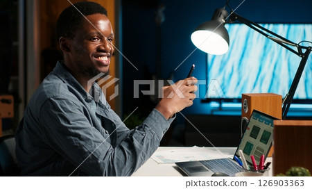 Independent adult teleworking from home and using his mobile phone, navigating social media and answering emails before starting his daily tasks completion. Comfortable modern setup. Camera B. 126903363